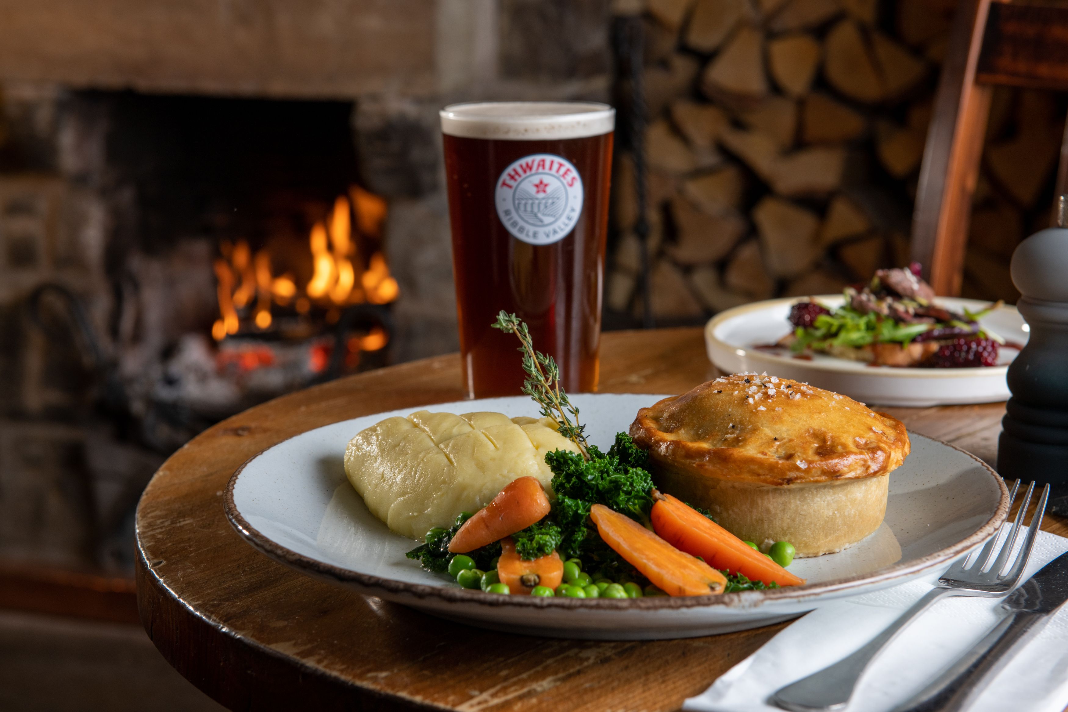 Pie, mash and vegetables on a plate with thwaites ale and fire in background