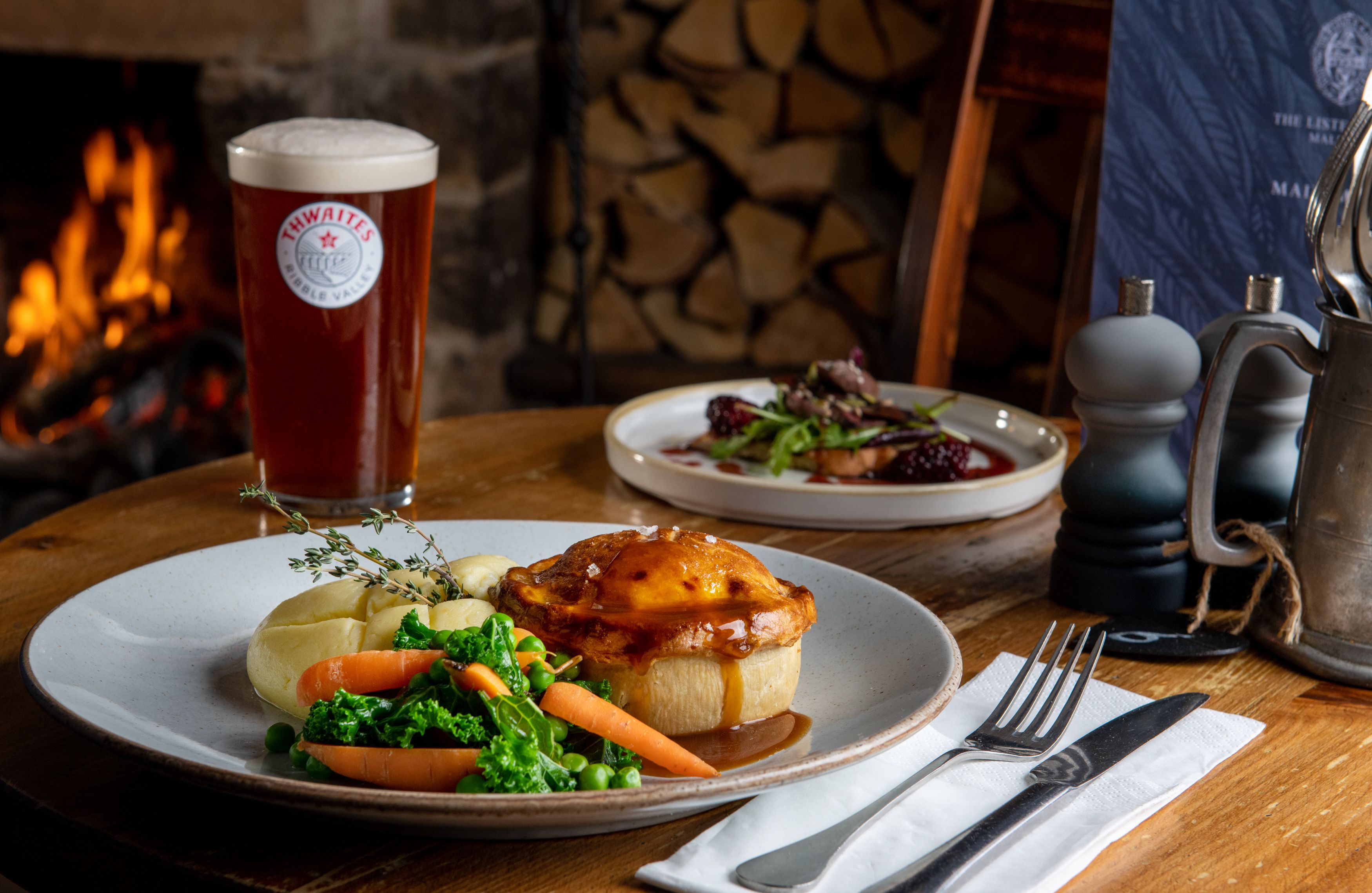 Pie, mash, vegetables and gravy on a plate with thwaites ale and fire in background