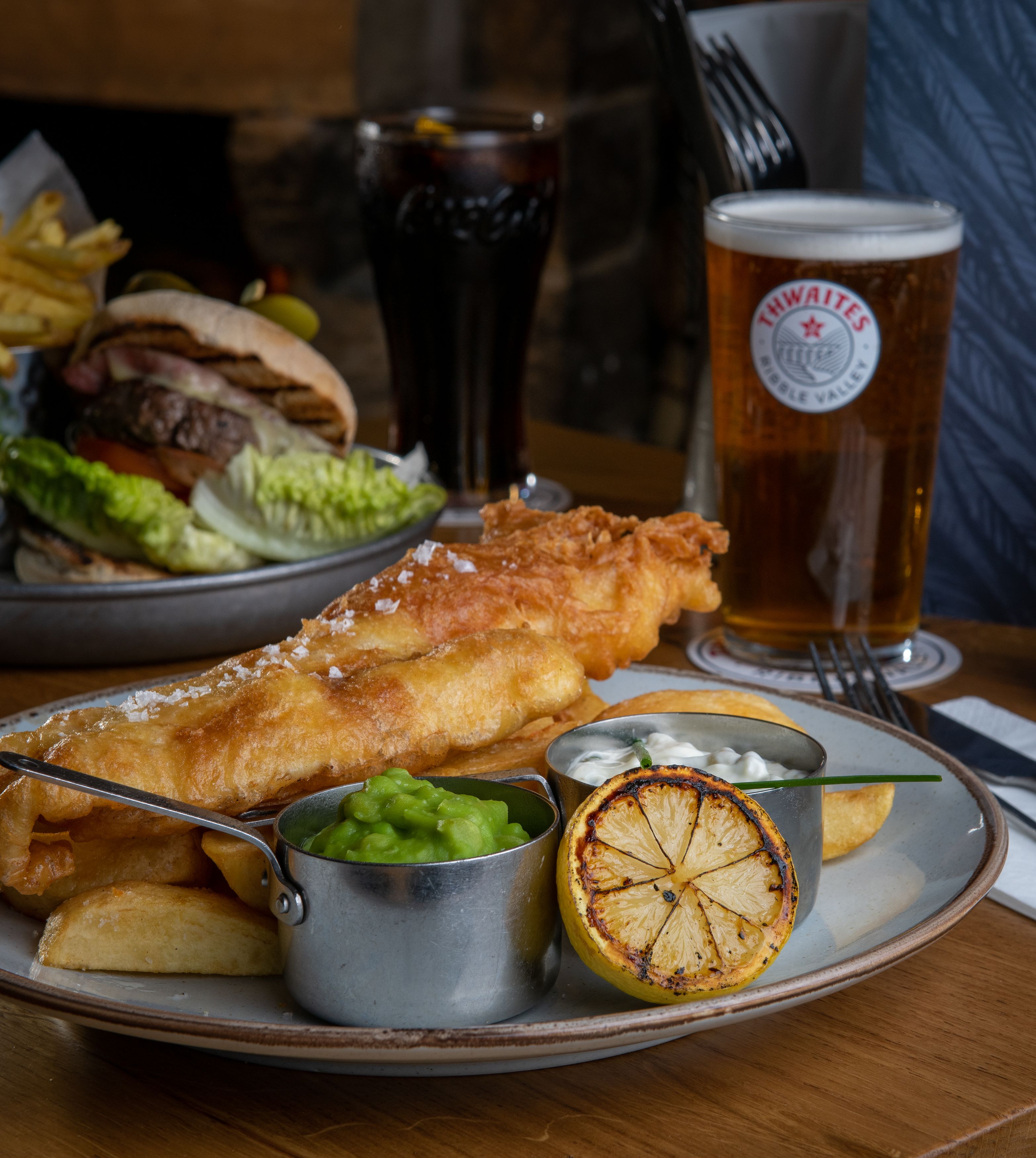 close up of fish and chips with mushy peas and thwaites ale and burger in background