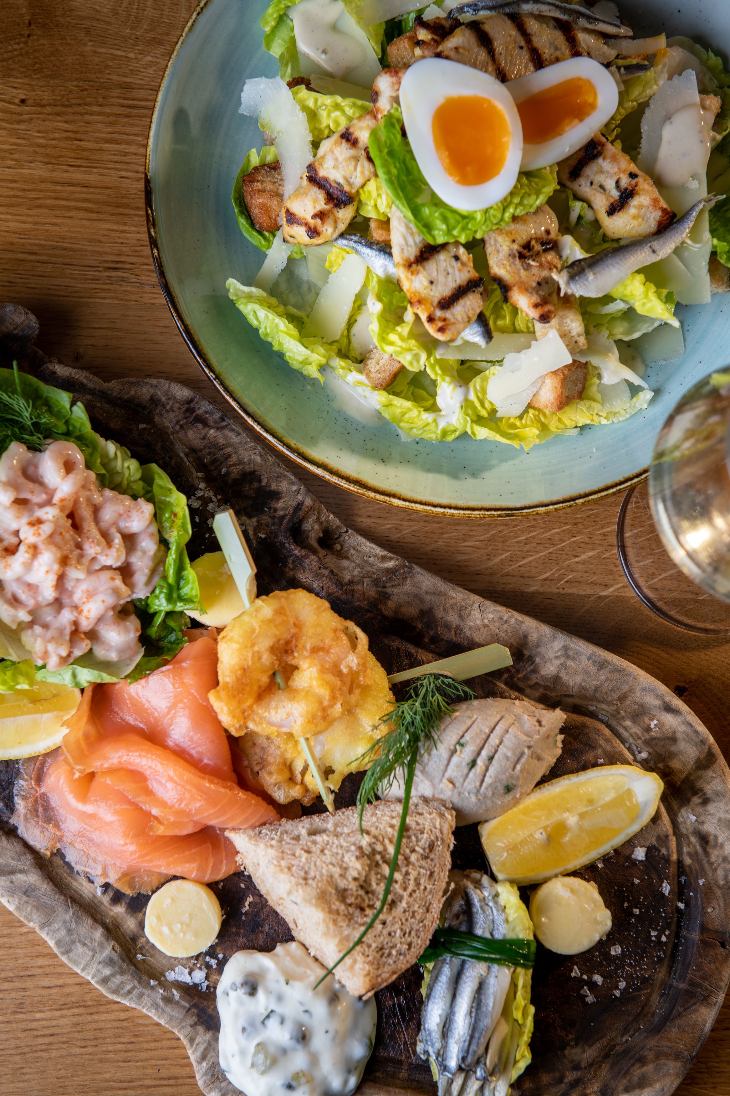 overhead shot of caesar salad with lettuce, chicken, egg and anchovies, and seafood platter on table